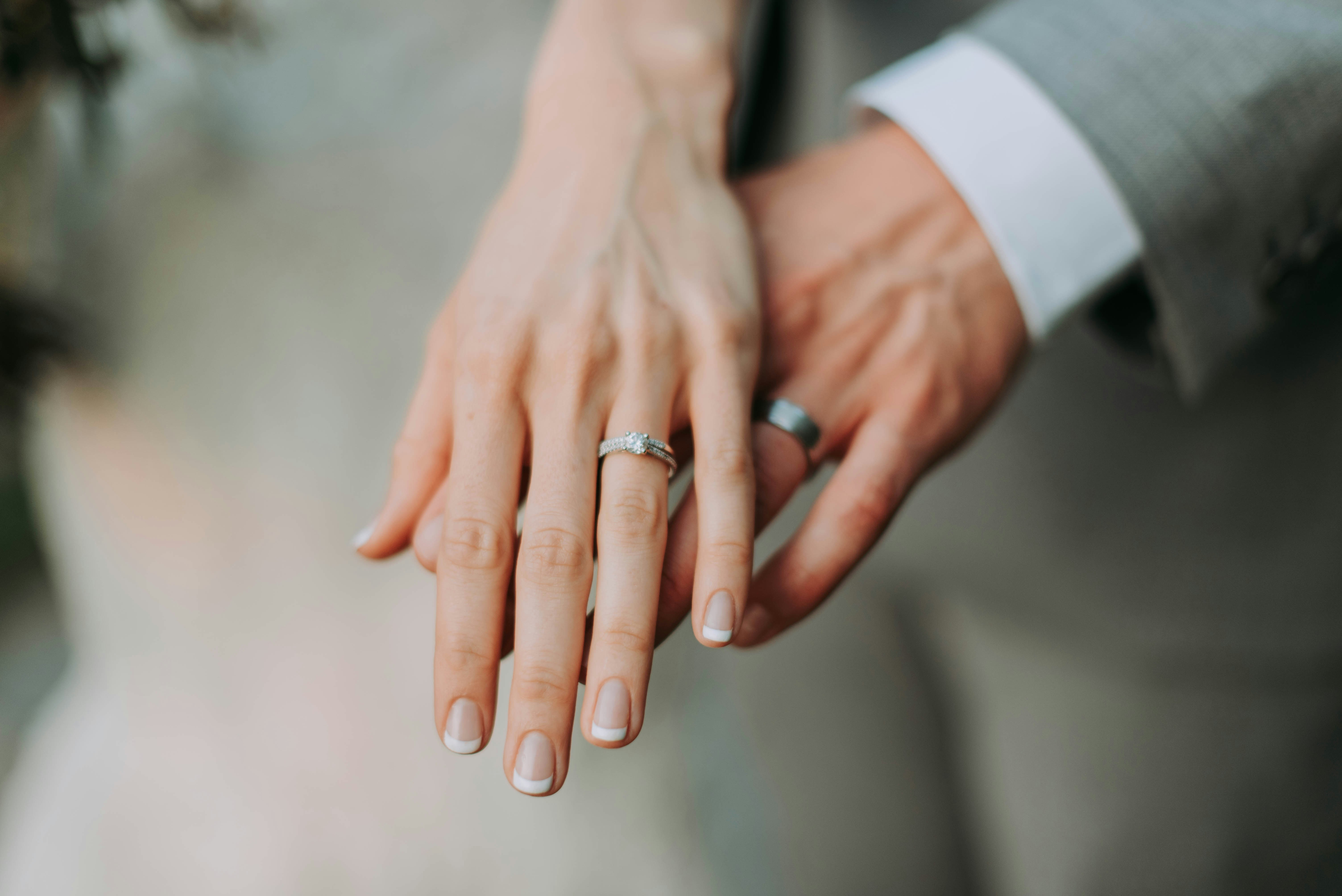 Close-up of two hands held together, showing a stone-set ring and a dark wedding band beside a white shirt cuff and grey suit sleeve.