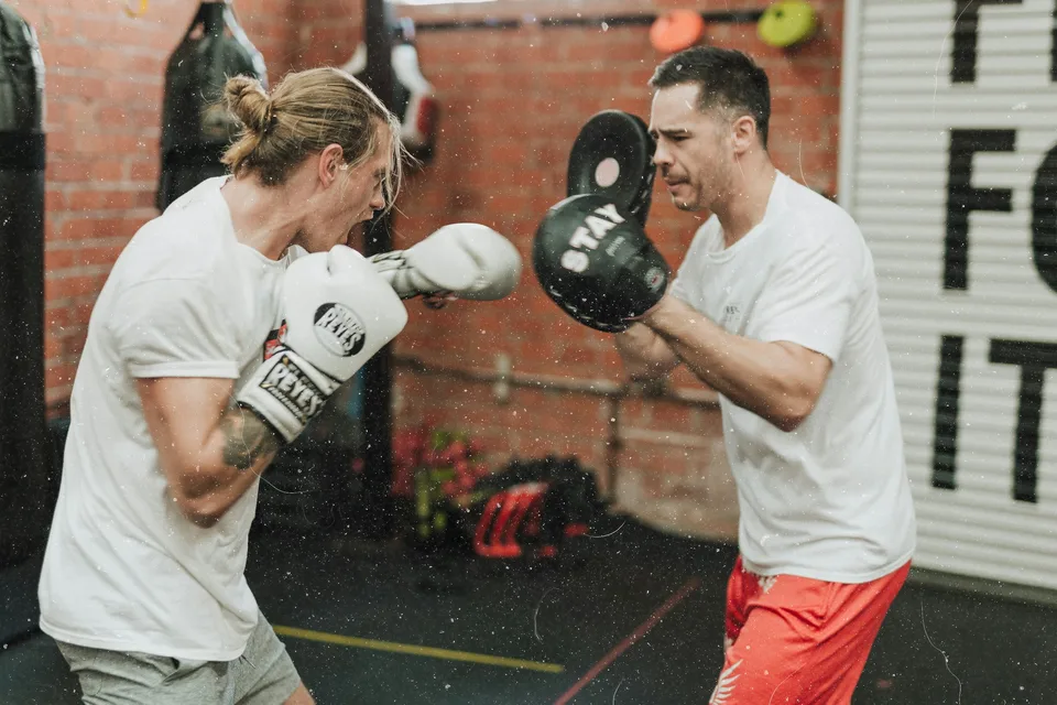 Two men in white T-shirts practising boxing pad work in a brick-walled gym.
