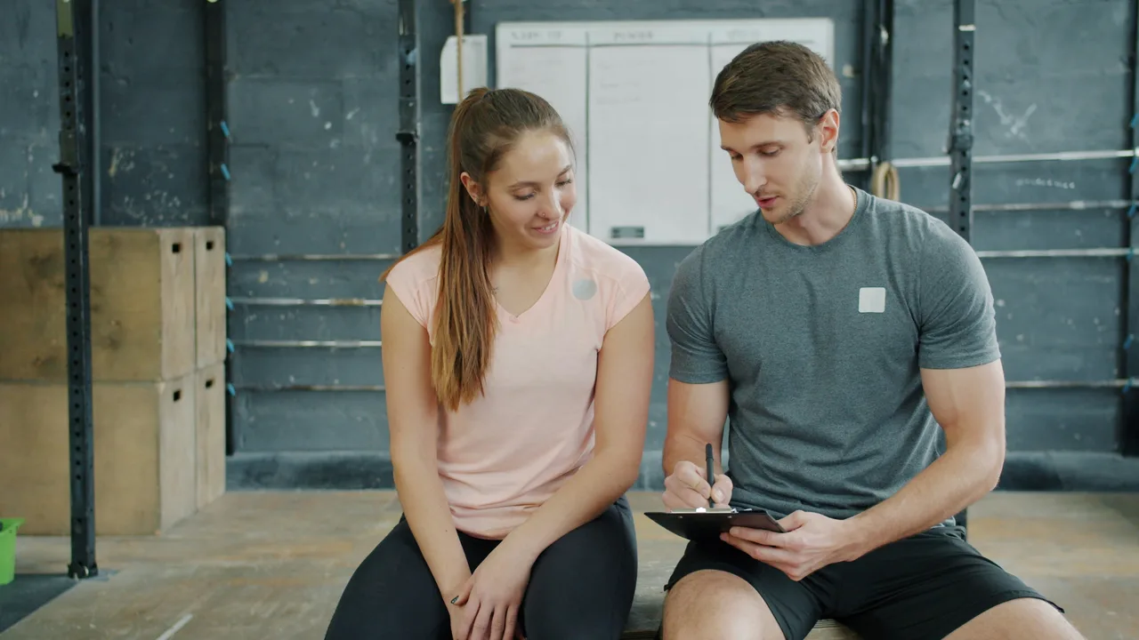 A coach writes on a clipboard while talking with a seated participant in a gym.