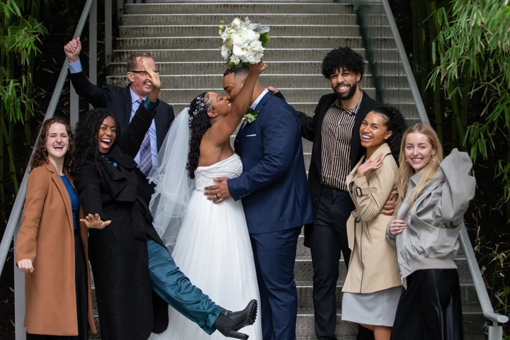 Bride and groom kissing at the foot of outdoor steps as a group of guests pose around them, cheering and lifting their arms.