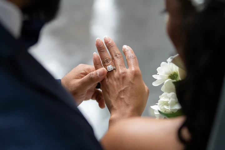 Close-up of the bride's raised hand and wedding ring as the groom touches her fingers, with her white dress softly blurred behind.