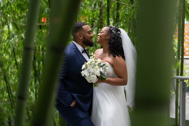 Bride and groom facing each other with a white bouquet between them, framed through thick green bamboo stems.