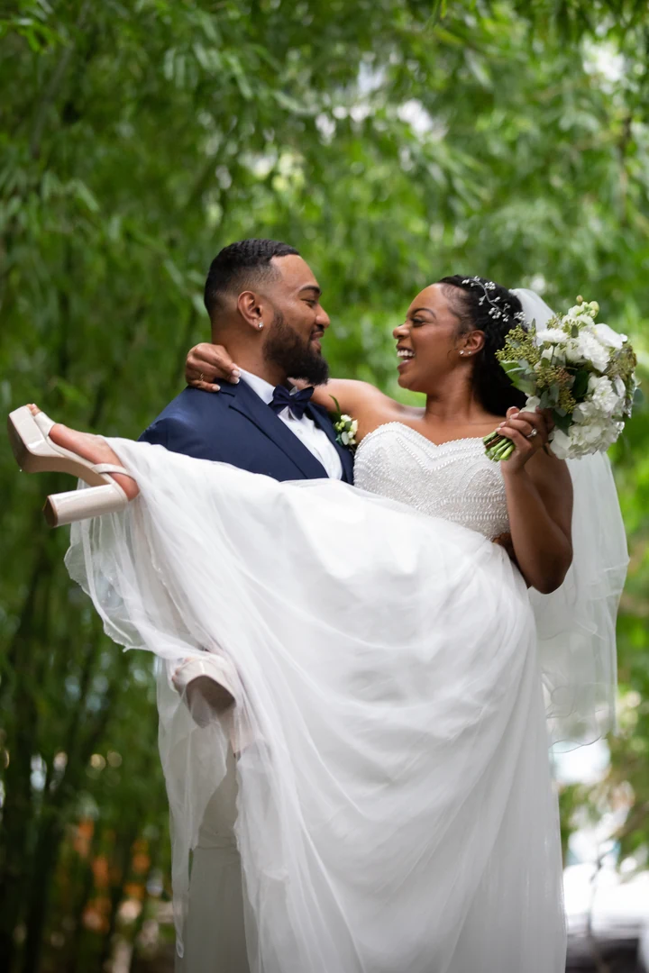 Groom carrying the bride in his arms among bamboo, as she smiles and holds a white bouquet beside her flowing dress.