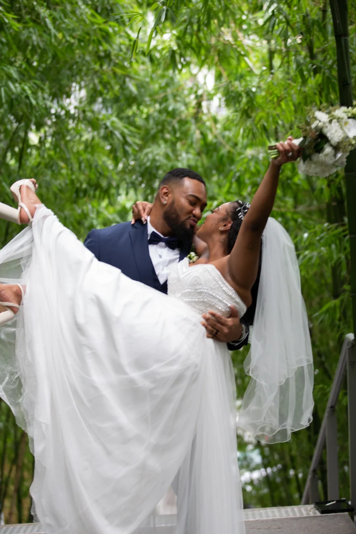 Groom carrying and kissing the bride near outdoor steps, as she raises her bouquet above them against green bamboo.