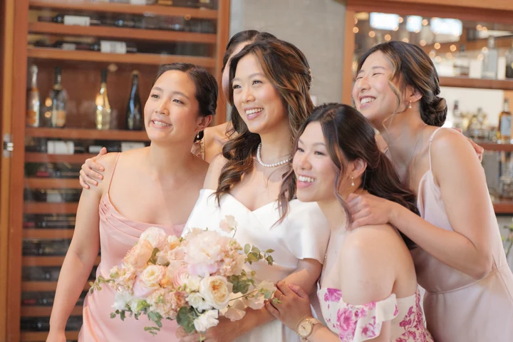 Bride and three bridesmaids smiling together indoors, holding pastel bouquets in front of wooden shelving.