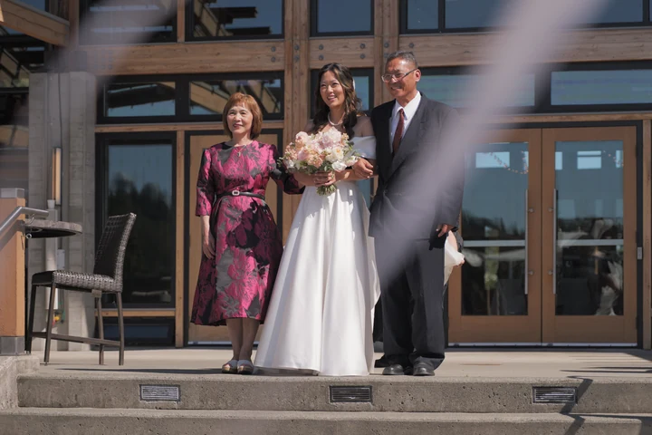 Bride holding a bouquet between a woman in a floral dress and a man in a dark suit on the steps outside a wood-and-glass building.