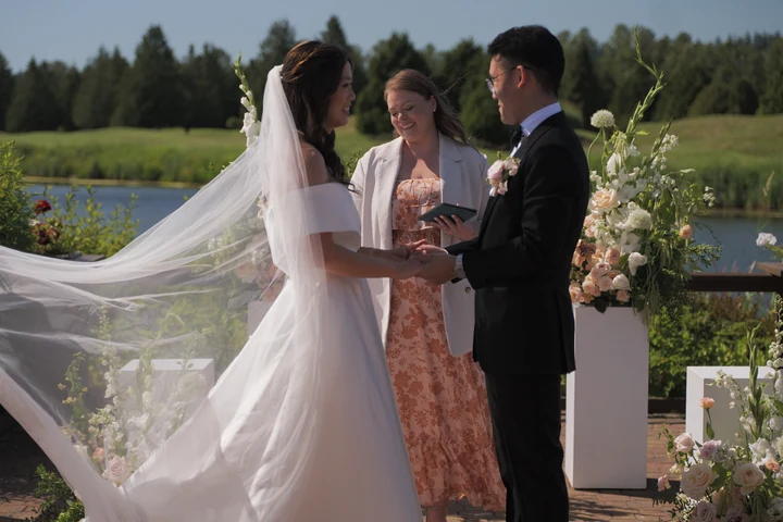 Bride and groom holding hands during an outdoor ceremony, with an officiant between them and the veil blowing toward the lawn.
