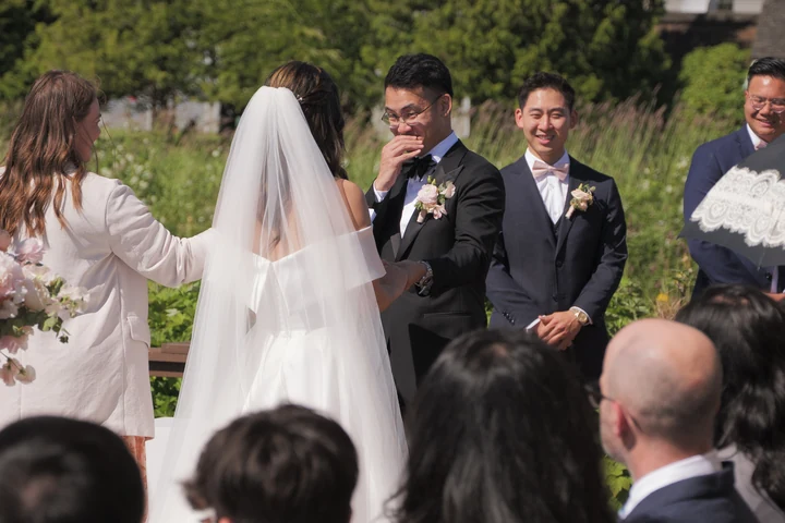 Groom wiping his eye as the veiled bride stands before him at an outdoor ceremony, with guests and attendants nearby.