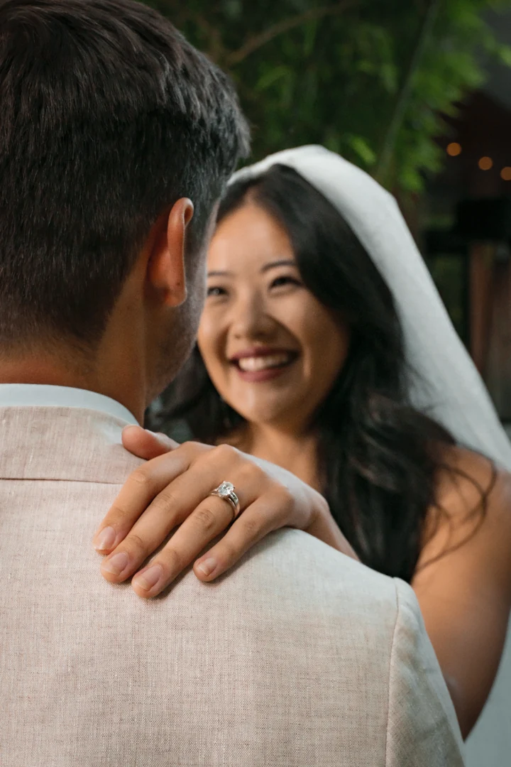 Bride smiling over the groom's shoulder, with her ring-bearing hand resting on his light-coloured suit and a veil framing her face.
