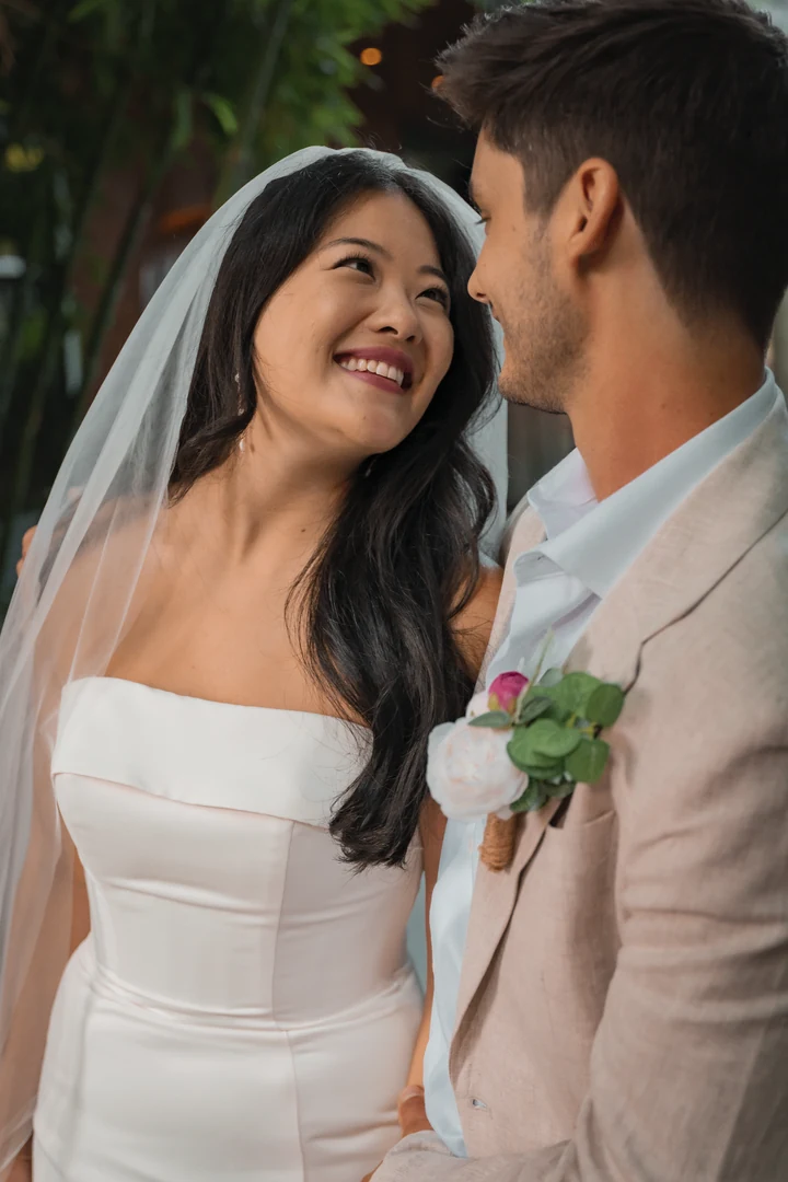 Bride smiling up at the groom in a light-coloured suit, with her veil, his floral boutonniere and green bamboo behind them.