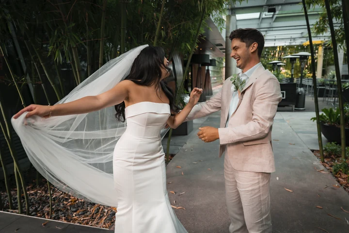 Bride turning beside the groom on a bamboo-lined walkway, holding her flowing veil outward as they smile at each other.