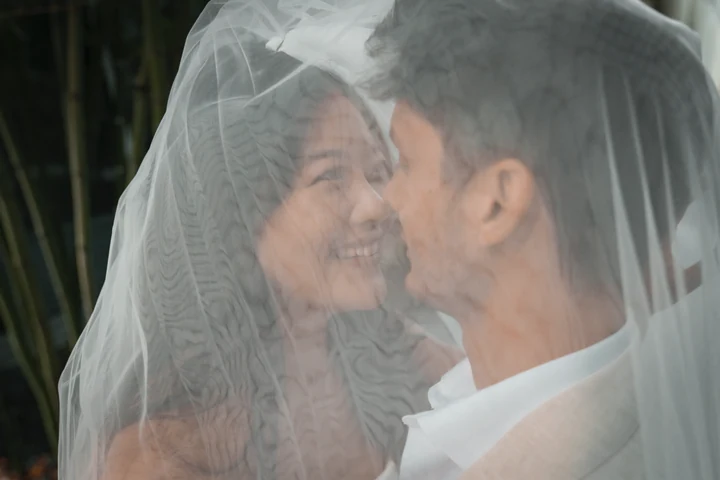Close-up of the bride and groom smiling face to face beneath a translucent veil, with their faces softly framed by the fabric.