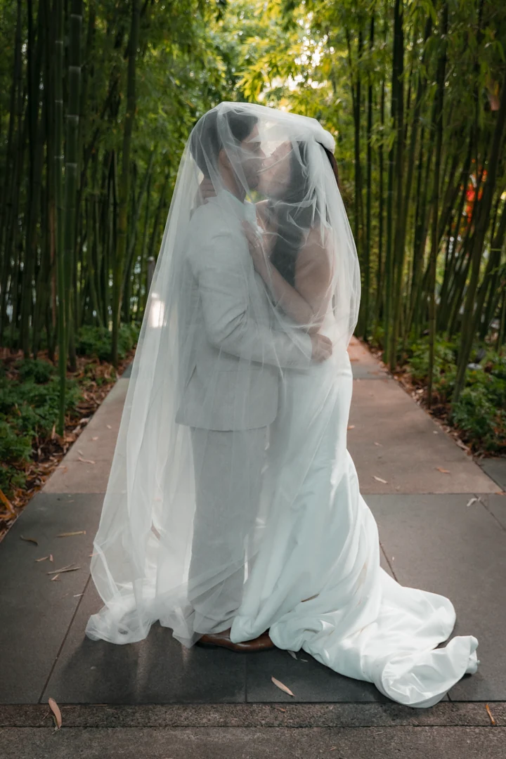 Full-length portrait of the bride and groom kissing beneath a veil on a narrow bamboo-lined path, with the dress train spread behind them.