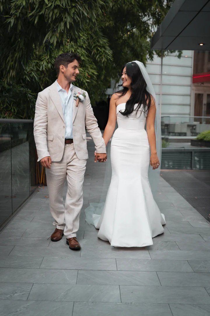 Bride and groom walking hand in hand along a paved walkway beside bamboo and glass walls, looking toward each other.