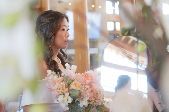 Bride holding a pastel pink and white bouquet beside a window, with her reflection visible in a round mirror.