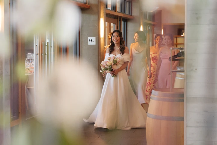 Bride in an off-the-shoulder white gown holding her bouquet inside a sunlit room, photographed through a softly blurred foreground.